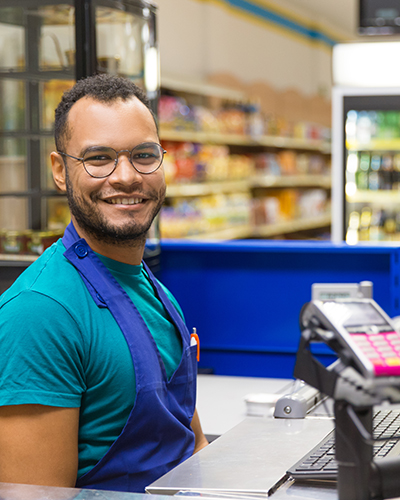 Smiling African American cashier sitting at checkout. Cheerful bearded young man in eyeglasses at workplace. Shopping concept