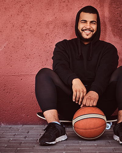 Cheerful African-American bearded guy dressed in a black hoodie and sports shorts sitting on a skateboard with basketball and leaning on wall.